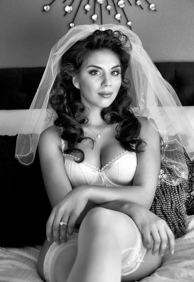 black and white portrait of a young woman wearing a white bra and panties and a wedding veil sitting on a bed in a bridal boudoir pose