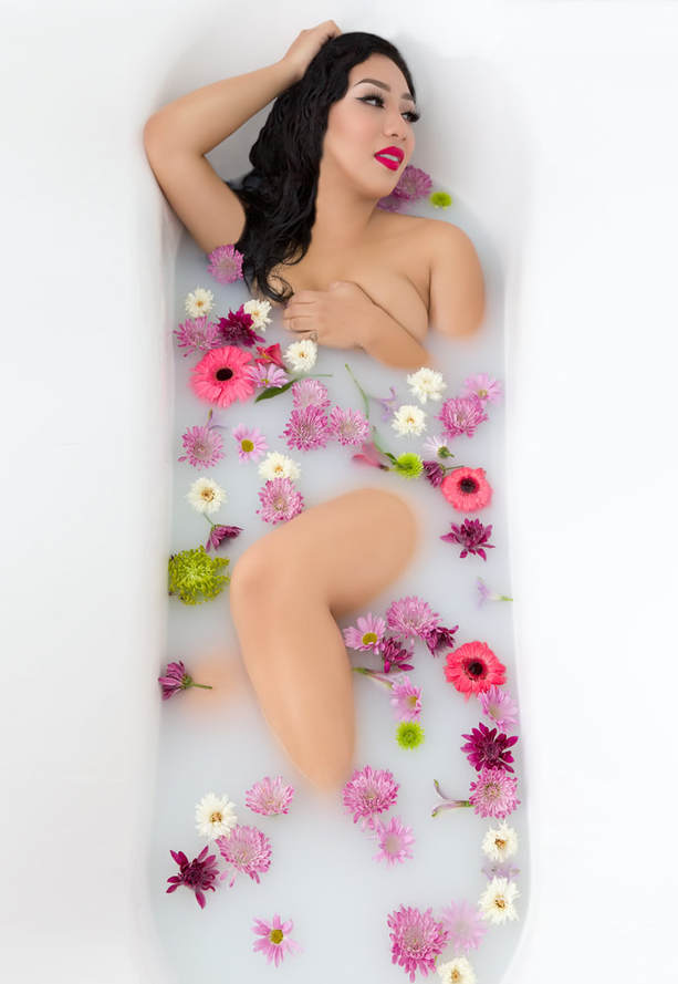 Photo of a beautiful young nude woman laying in a milk bath with flowers surrounding body during a boudoir photo shoot.