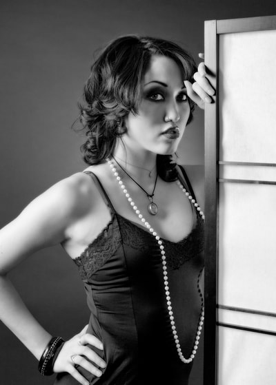 Black and white glamour photo of a young woman wearing a black dress posing next to a changing screen